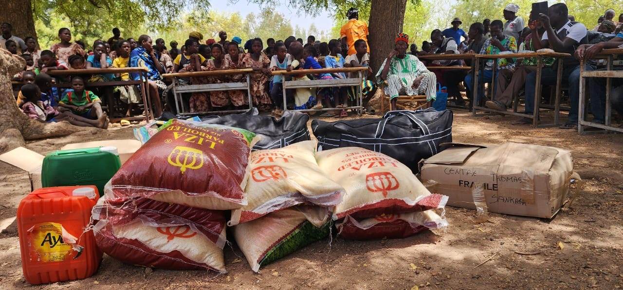 Denrées et matériel rassemblés en vue de leur remise à la communauté de Ménéga, avec les habitants réunis à l'arrière-plan, lors de la mission ICEV, Burkina Faso, avril 2026.
